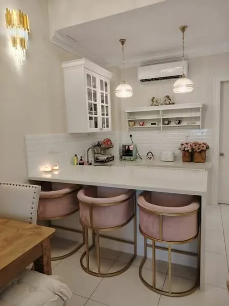 Kitchen features: white cabinetry with glass-front upper cabinets, white tiled backsplash, marble countertop, 3 pink upholstered bar stools with gold frames, built-in sink, chrome faucet, recessed downlights, wall-mounted air conditioning unit, decorative shelf with small items, table with candles, white ceramic teapot, pink flowers in vase; style: modern.