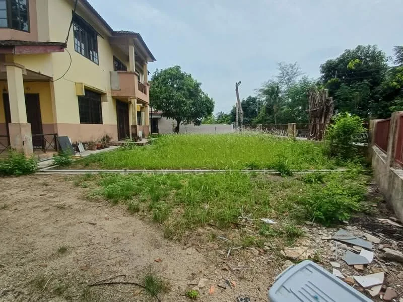 Exterior type: semi-detached house; features: two-storey building, unmaintained garden area, overgrown grass, surrounding trees, concrete pathway, fenced perimeter, adjacent structures, dirt ground, partially visible exterior walls, balcony on upper floor; style: traditional.