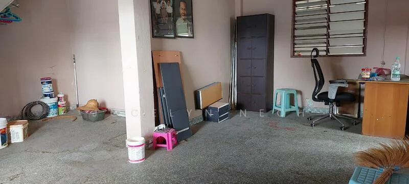 Room features: concrete flooring, painted walls in beige, window with horizontal slats, desk with wooden surface, office chair, storage cabinet, various paint cans, plastic stools in green and pink, broom, wire basket.