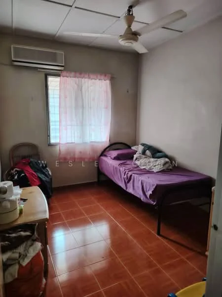 Bedroom features: single bed with purple bedding, wooden headboard, ceiling fan, wall-mounted air conditioning unit, window with sheer curtains, brown tiled flooring, wooden chair, small table, clothes on the floor.