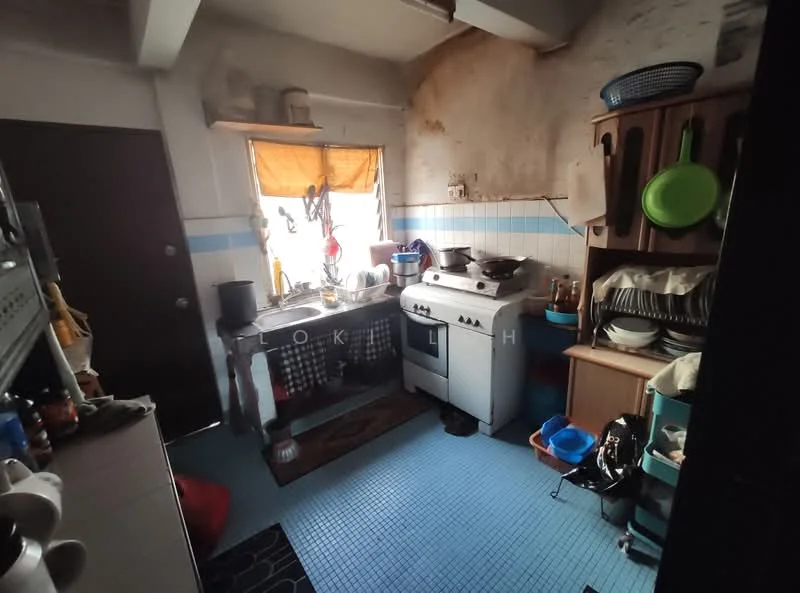 Kitchen features: white walls with peeling paint, blue tiled backsplash, blue vinyl flooring, single-bowl stainless-steel sink, chrome faucet, freestanding gas stove, wooden cabinet, open shelving, various cooking utensils, stacked plates and bowls, window with orange curtain, kitchen towel hanging, black rug, kitchen mat.