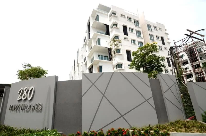 Exterior type: low-rise apartment block; features: white painted facade, glass balconies, concrete structure, landscaped garden, manicured flower beds, modern architectural design, prominent signage, surrounded by greenery; style: modern.