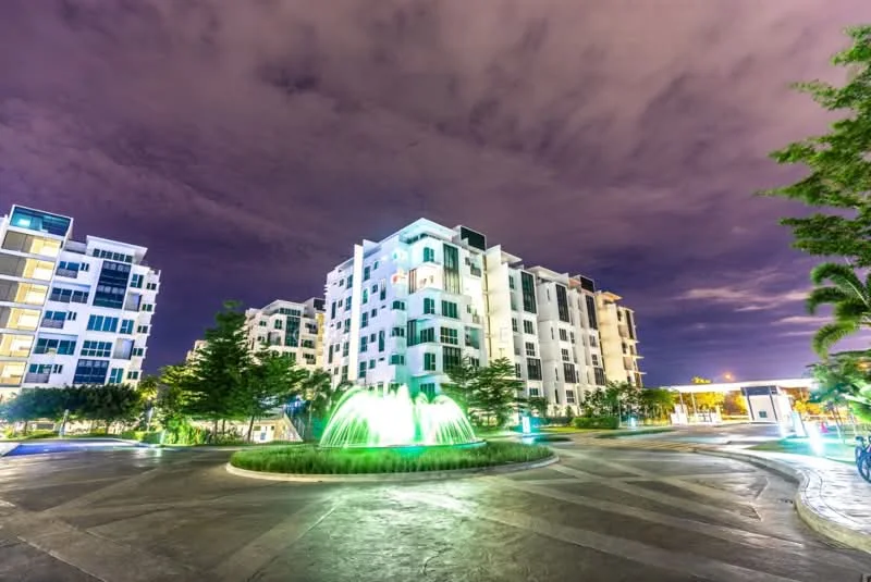 Exterior type: low-rise apartment block; features: white painted facade, glass windows, landscaped garden with fountain, manicured lawn, paved driveway, outdoor lighting, surrounded by trees, multiple storeys; style: modern.