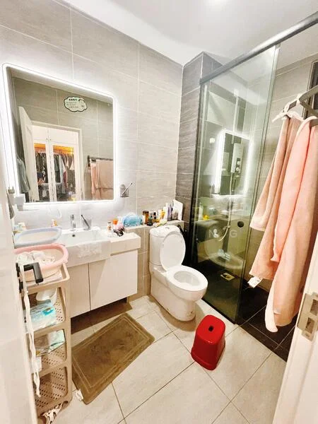 Bathroom features: single-sink vanity with white cabinet, framed mirror with LED lighting, toilet, walk-in shower with glass enclosure, chrome basin mixer, grey wall tiles, light grey floor tiles, towel rack, shower mat, storage cart, various toiletries on countertop, pink bath towels hanging; style: modern.