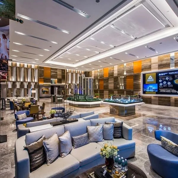 Living features: sofa with light blue upholstery, decorative cushions, coffee table, marble flooring, recessed downlights, feature wall with wood paneling, large indoor plants, artificial water feature, modern lighting fixtures, wall-mounted screens, decorative vases, open layout, high ceiling, glass partitions, seating area with armchairs; style: modern.