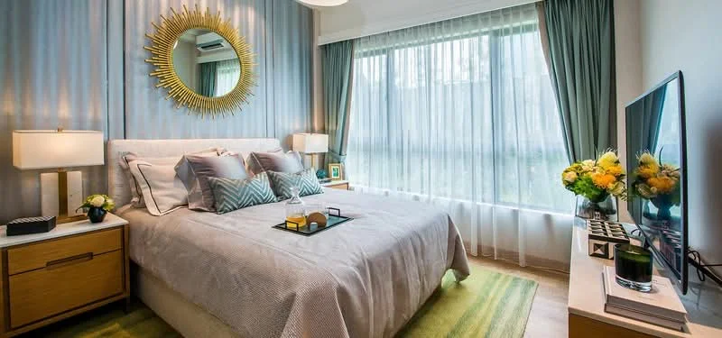 Bedroom features: queen-size bed with upholstered headboard, decorative throw pillows, bedside tables with lamps, wall-mounted TV, green area rug, floor-to-ceiling windows with sheer curtains, gold sunburst mirror, wooden dresser, potted flowers on dresser, air conditioning unit, grey accent wall, white painted walls, recessed ceiling lights; style: modern.