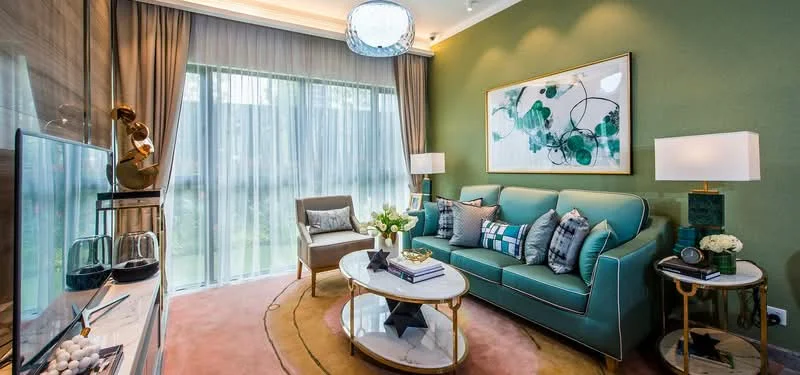 Living features: green accent wall, light grey curtains, floor-to-ceiling windows, 3-seater teal sofa, decorative cushions in various patterns, round coffee table with gold trim, side table with lamp, framed artwork on wall, TV console with decorative items, area rug with circular pattern, armchair in light grey fabric, decorative vase with flowers; style: modern; view: garden.
