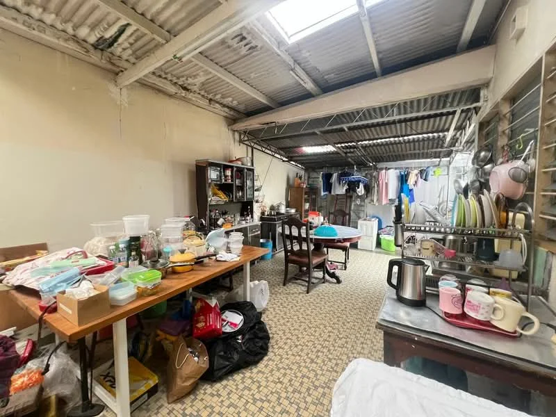 Laundry features: metal roof, exposed beams, tile flooring, wooden dining table, plastic storage containers, dish rack with dishes, kettle, various kitchenware, clothes drying rack, washing machine, clothes hanging on line, plastic bags, shelves with storage items, light source from skylight.