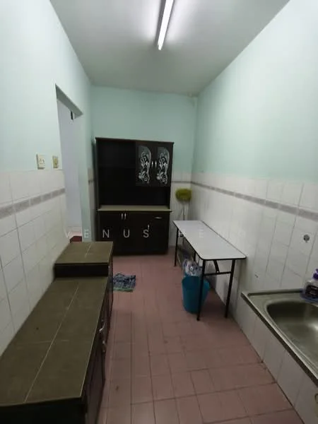 Kitchen features: light blue painted walls, white tiled backsplash, pink tiled flooring, brown wooden cabinets, countertop with dark surface, metal sink, single-bowl stainless-steel sink, overhead fluorescent lighting, table with metal legs, plastic trash bin, cleaning cloth on floor.