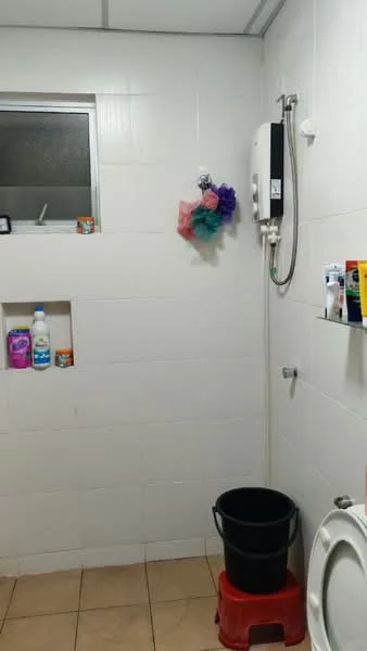 Bathroom features: white tiled walls, cream tiled flooring, shower with wall-mounted water heater, toilet, bucket, red stool, shelf with toiletries, niche for storage, window for natural light.