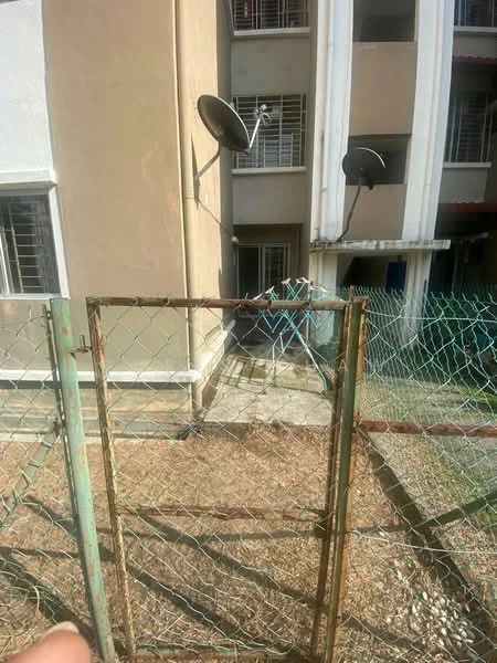 Exterior type: low-rise apartment block; features: fenced yard with chain-link fence, two satellite dishes mounted on the building, exterior wall with beige paint, ground level access, visible windows with metal grills, concrete walkway leading to the entrance; style: modern.