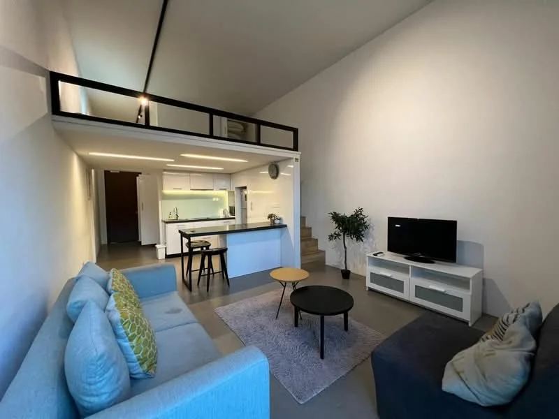 Living features: grey fabric sofa, light blue fabric sofa with decorative cushions, round black coffee table, area rug with geometric pattern, wall-mounted TV, TV console with storage, open-plan layout, high ceiling, staircase leading to loft area, greenery in ceramic pot, recessed lighting, kitchen island with seating, white cabinetry, modern appliances; style: modern.