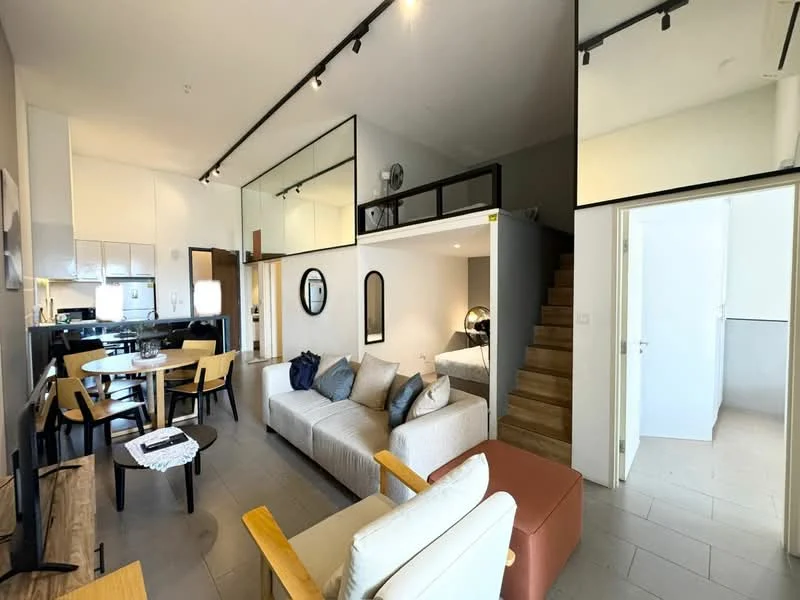 Living features: sofa in light grey fabric, wooden coffee table, TV console, wall-mounted TV, pendant light, open shelving, decorative mirror, area rug, ceiling fan, wooden staircase, large windows, dining table with chairs, kitchenette with white cabinets, black countertop, stainless-steel sink; style: modern. Room features: white walls, tile flooring, glass partition, wooden staircase.