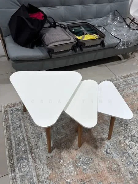Room features: triangular coffee table with white surface, wooden legs, grey upholstered sofa, decorative area rug with intricate pattern, luggage and bags on sofa, light-colored tiled flooring; style: modern.