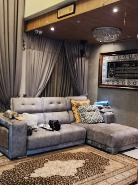 Living features: L-shape sectional sofa in grey, decorative cushions in yellow and patterned fabric, area rug with intricate pattern, wall-mounted artwork with Arabic script, chandelier with crystals, floor-to-ceiling curtains in grey, wood paneling on ceiling, recessed lighting, coffee table, decorative items on the sofa; style: contemporary.