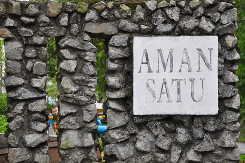 Exterior type: main entrance gate; features: stone cladding, decorative stone wall, signage with text 'AMAN SATU', surrounding greenery; style: modern.