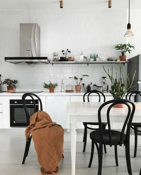 Kitchen features: white tiled backsplash, stainless-steel range hood, built-in oven, freestanding refrigerator, white cabinetry, open shelving, black dining chairs, white dining table, indoor plants in pots, pendant lighting, wooden flooring, textured brown throw, glass containers on shelf, kitchen utensils on display; style: modern.