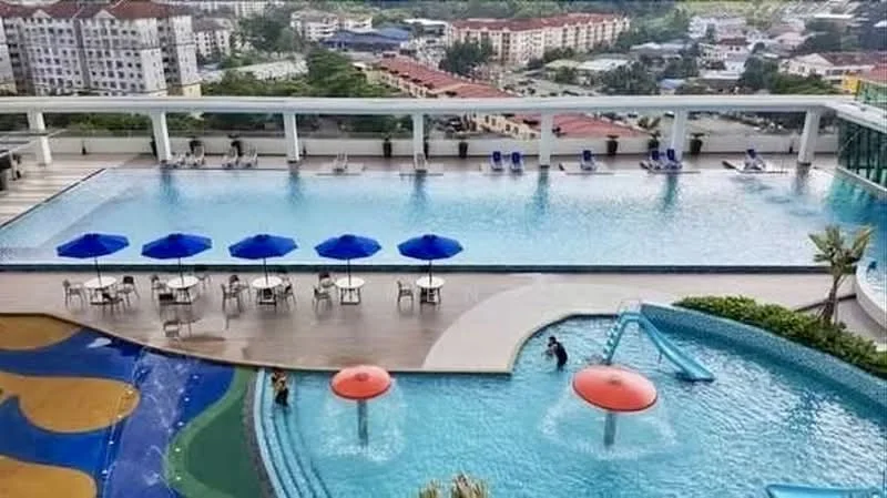 Exterior type: swimming pool deck; features: infinity pool, children's splash pool, pool deck with loungers, water slide, umbrellas over seating areas, landscaped greenery around pool, surrounding residential buildings, view of city skyline; style: modern.