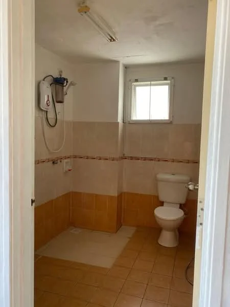 Bathroom features: white toilet, shower area with tiled walls, tan ceramic tile flooring, white wall-mounted water heater, small window for natural light, light fixture on ceiling, tiled backsplash, grout lines visible.