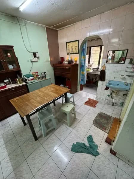 Kitchen features: wooden dining table, 2 plastic stools, green painted walls, white tiled walls, white tiled flooring, sink with faucet, kitchen cabinets, countertop with appliances, window with curtain, decorative wall art, floor mat, cleaning cloth on floor.