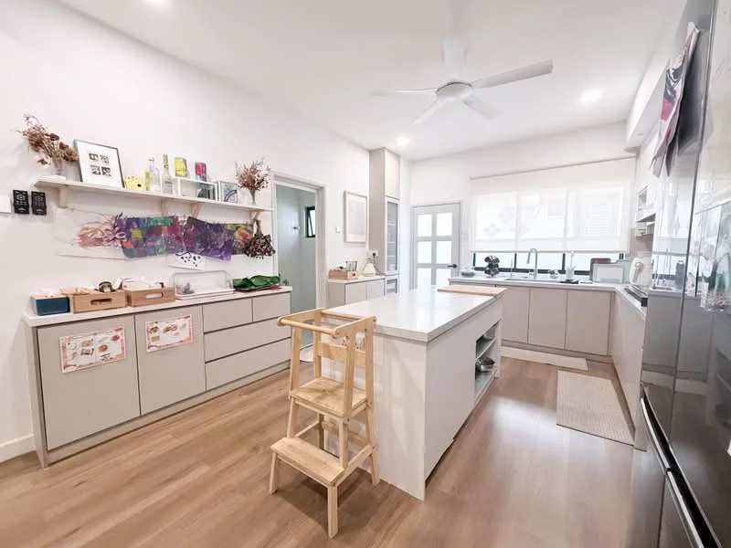 Kitchen features: white painted walls, light wood flooring, island countertop with seating, stainless-steel sink, chrome pull-down faucet, built-in oven, freestanding range oven, white cabinetry with flat-panel doors, open shelving, recessed downlights, 5-blade ceiling fan, window with natural light, decorative wall art; style: modern.