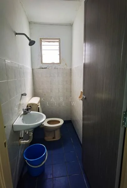 Bathroom features: white tiled walls, blue ceramic tile flooring, single-sink vanity, toilet, shower with wall-mounted shower head, small window, wooden door, bucket.