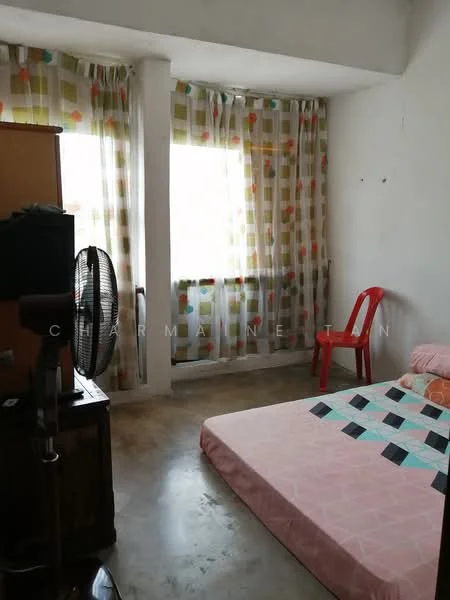 Bedroom features: concrete flooring, white painted walls, floor-to-ceiling windows, sheer curtains with colorful pattern, single bed with pink bedding, wooden wardrobe, red plastic chair, table fan, decorative throw pillows.