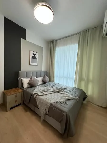 Bedroom features: queen-size bed with grey upholstered headboard, two decorative pillows, light grey throw blanket, nightstand with wooden top and drawer, framed artwork on wall, floor-to-ceiling windows with sheer curtains, light green curtains, wooden flooring, ceiling light fixture, wall-mounted split AC; style: modern.