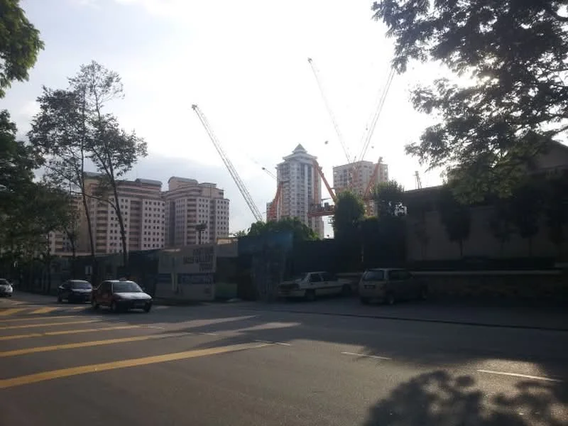Exterior type: high-rise condominium; features: multiple construction cranes, surrounding trees, urban street view, residential buildings in background, partially visible building facade, road with yellow lane markings; style: modern.
