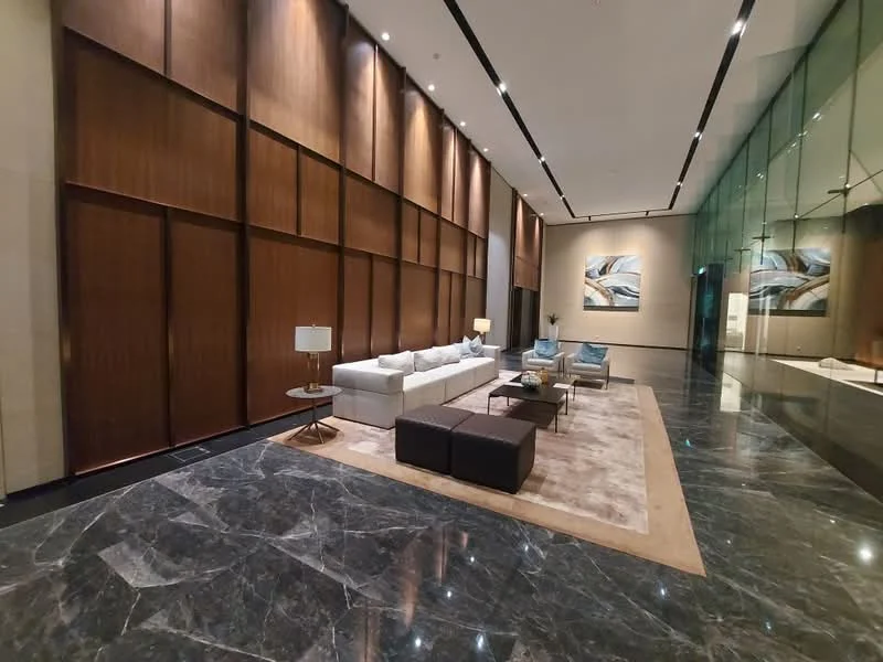 Living features: grey marble flooring, wood panelled accent wall, white fabric sofa, two blue upholstered armchairs, coffee table with dark wood finish, area rug with neutral tones, table lamp with white shade, framed artwork on wall, recessed LED downlights, large glass windows, high ceiling; style: modern.