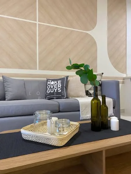 Living features: grey fabric sofa, decorative throw pillow with text, wood coffee table, glass vase with greenery, two glass bottles, white candle, woven tray, textured wall paneling, neutral wall color; style: modern.