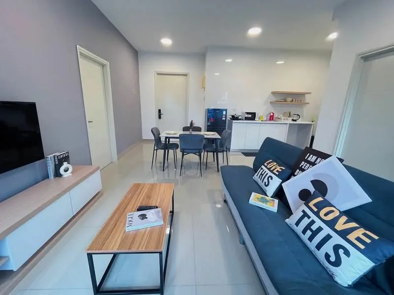 Living features: grey fabric sofa, coffee table with wooden top and black metal frame, wall-mounted TV, light grey painted walls, recessed downlights, decorative cushions with text, open shelving unit, dining table with four chairs, kitchen area visible in background, white cabinetry, stainless-steel fridge, countertop with kitchen appliances; style: modern.
