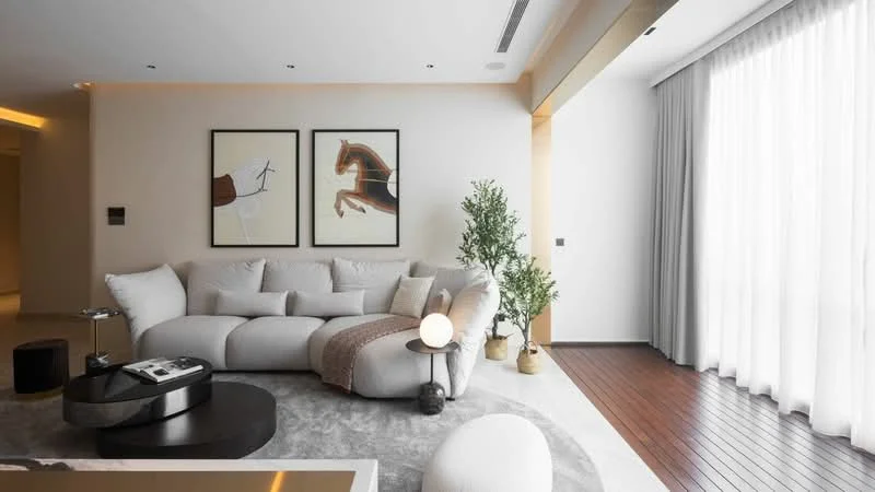 Living features: L-shape sectional sofa in light grey fabric, round coffee table with black surface, area rug with circular shape, framed artwork on walls, floor-to-ceiling windows with sheer curtains, wooden flooring, decorative indoor plant, table lamp with round base; style: modern.
