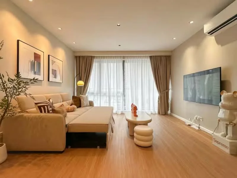 Living features: L-shape sectional sofa in beige, coffee table with rounded design, wall-mounted TV, floor-to-ceiling windows, sheer curtains, wooden flooring, recessed downlights, decorative plant, decorative sculpture, wall art with abstract designs, air conditioning unit; style: modern.