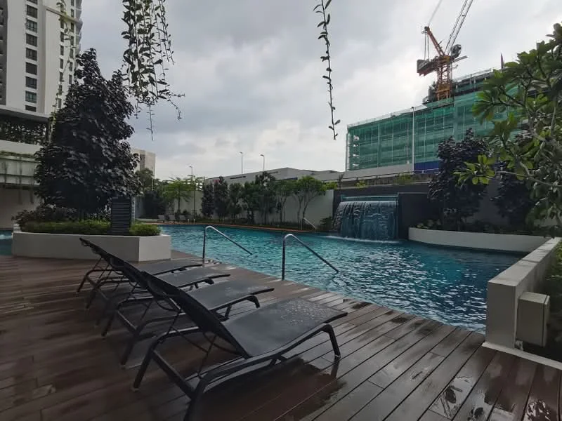 Exterior type: swimming pool deck; features: infinity pool, waterfall feature, wooden deck flooring, poolside loungers, landscaped greenery, tropical plants, surrounding construction site, overcast sky; style: modern.