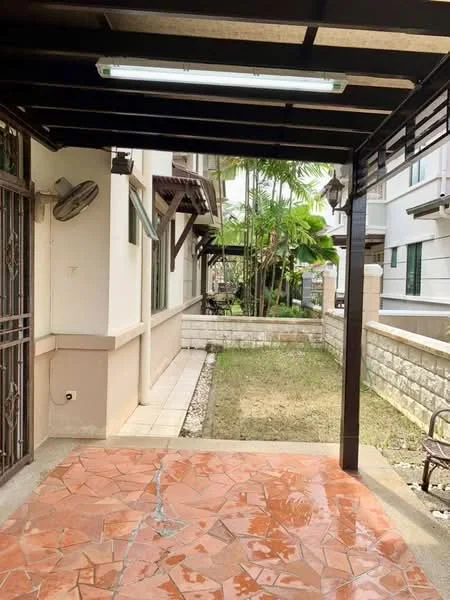 Exterior type: linked terrace house; features: covered porch with metal roof, terracotta tile flooring, stone boundary wall, manicured garden area, paved walkway, exterior wall with white paint, window with wooden shutters, visible palm trees in garden; style: modern.