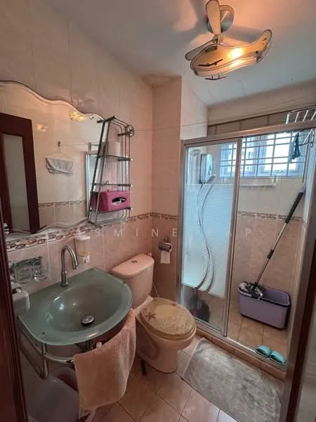 Bathroom features: glass shower enclosure, chrome handheld shower head, toilet, single-sink vanity with glass basin, chrome faucet, mirror with decorative frame, towel rack, tile flooring, beige wall tiles, light fixture with ceiling fan, shower mat, cleaning supplies visible; style: modern.