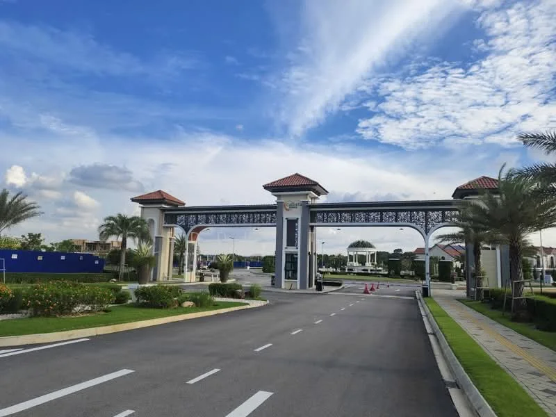 Exterior type: main entrance gate; features: ornate entrance gate with decorative metalwork, two towers with red tiled roofs, paved driveway, landscaped garden with flowering plants, palm trees lining the entrance, open-air pavilion visible in the background, blue sky with scattered clouds; style: modern.