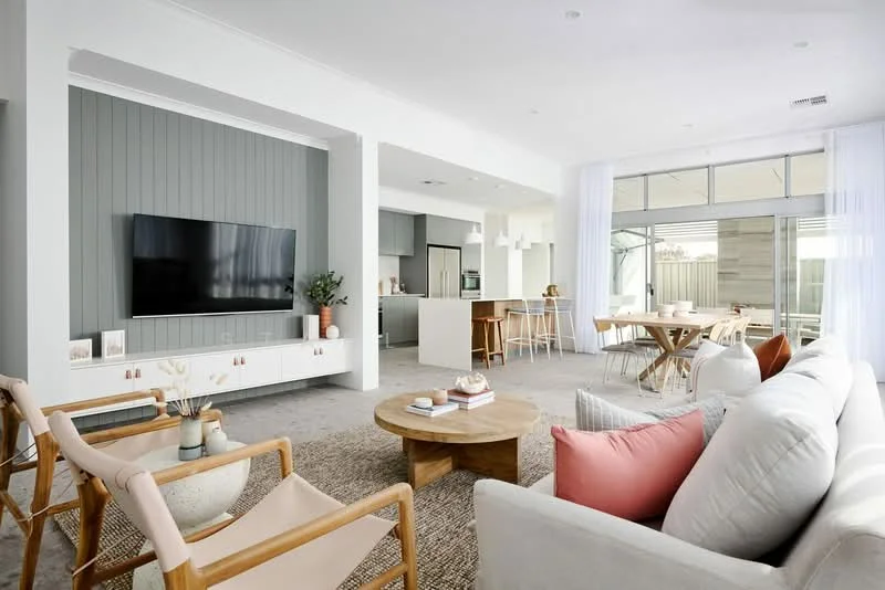 Living features: light grey walls, accent wall with vertical wood paneling in grey, large wall-mounted flat-screen TV, light beige area rug, 3-seater fabric sofa in white, wooden coffee table, 2 wooden armchairs with light fabric, decorative cushions in pink and grey, large windows with sheer curtains, open layout to dining area and kitchen; style: modern; view: garden. Dining features: round wooden dining table, 4 wooden dining chairs, large sliding glass door, natural light from windows; style: modern; view: garden. Kitchen features: open kitchen layout, island counter with bar stools, modern cabinetry, stainless-steel appliances; style: modern; view: garden.