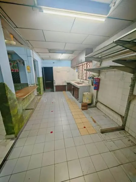Kitchen features: white tiled walls, green painted walls, grey tiled flooring, wooden countertop, sink, stainless-steel faucet, wall-mounted shelving, overhead fluorescent lighting, open layout, window with metal grilles, kitchen cabinets, space for appliances.