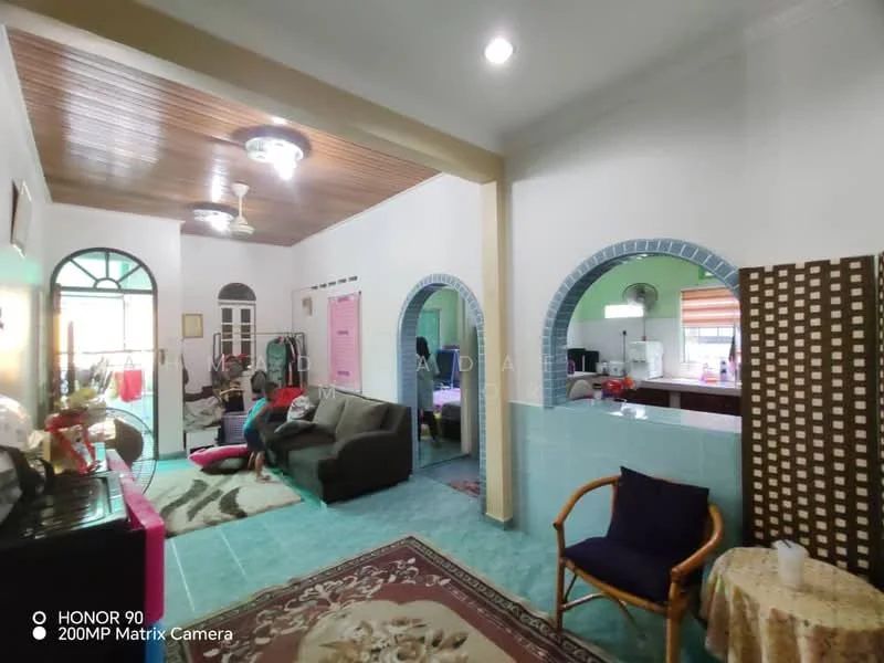 Living features: green ceramic tile flooring, white painted walls, wooden ceiling with exposed beams, 5-blade ceiling fan with light, sofa in grey fabric, coffee table, area rug with floral pattern, wall-mounted TV, decorative wall with arches, window with sheer curtains; style: traditional. Kitchen features: green ceramic tile flooring, white painted walls, kitchen counter with cabinets, window with blinds, refrigerator, stove, sink, open layout to living area; style: traditional.