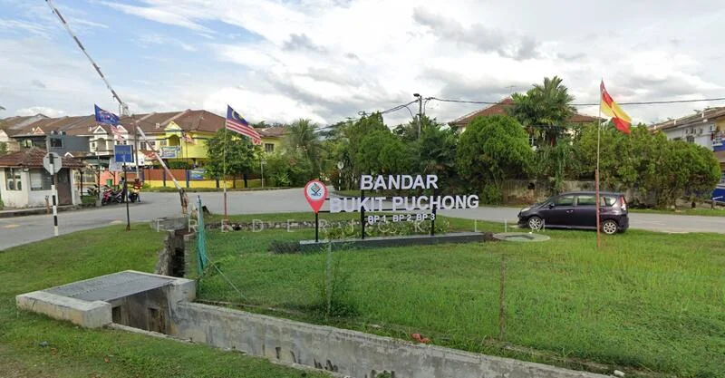 Exterior features: signage with 'BANDAR BUKIT PUCHONG', manicured lawn, surrounding greenery, paved road, residential buildings in background, flag poles with flags, car parked on roadside; style: modern.