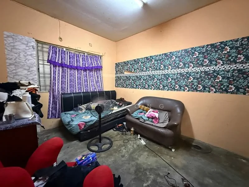 Room features: orange painted walls, floral wallpaper, concrete flooring, single bed with mattress, brown leather sofa, ceiling fan, curtains with purple pattern, wooden desk, cluttered space.
