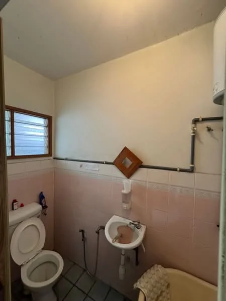Bathroom features: white toilet, small sink, mirror, light fixture, pink tiled walls, grey tiled flooring, window, water heater.