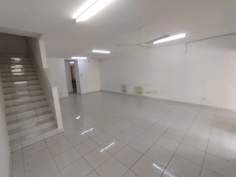 Room features: white painted walls, glossy white tile flooring, ceiling with fluorescent lighting, ceiling fan, staircase leading to upper level, open layout, no visible furniture, large empty space; style: minimalist.