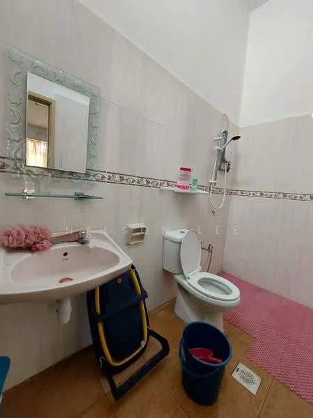 Bathroom features: single-sink vanity with wall-mounted mirror, chrome basin mixer faucet, toilet, shower with handheld showerhead, tile walls in light color, tile flooring, pink bath mat, plastic bucket, stool, shelf with toiletries.