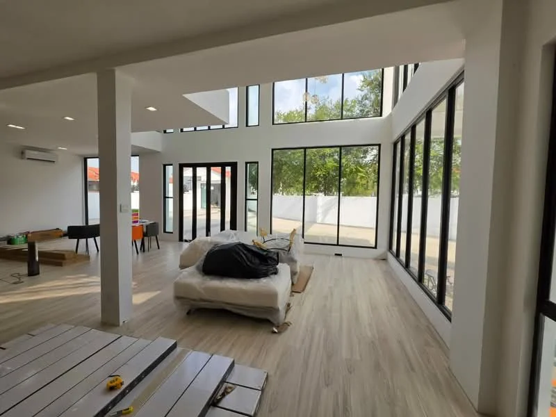 Living features: large floor-to-ceiling windows, white painted walls, light wood flooring, exposed ceiling beams, recessed downlights, air conditioning unit, sofa with black throw, coffee table, open layout, natural light from multiple angles, view of outdoor space; style: modern; view: garden.