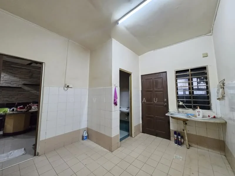 Room features: white tiled walls, light beige tiled flooring, single-bowl stainless-steel sink, wooden cabinet, window with metal bars, fluorescent ceiling light, door leading to another room, corner with plumbing fixtures, hanging towel rack, wall-mounted soap dispenser.