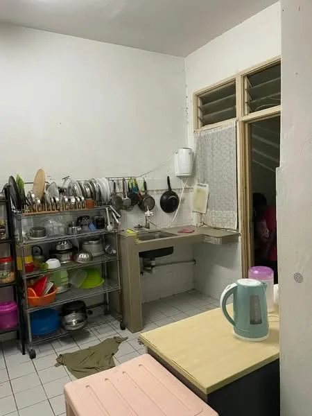 Kitchen features: white painted walls, tile flooring, stainless-steel sink, chrome faucet, kitchen shelving unit with pots and pans, various colored plastic bowls, kettle, countertop, pink storage box, window with sheer curtain, open layout to adjacent room.