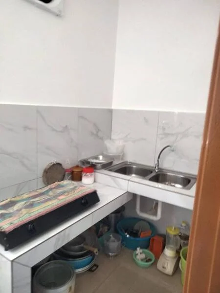 Kitchen features: double-bowl stainless-steel sink, chrome faucet, white marble backsplash, white tiled walls, light-colored floor tiles, countertop with storage underneath, gas stove, various kitchen utensils, blender, plastic containers, dish rack, cleaning supplies.